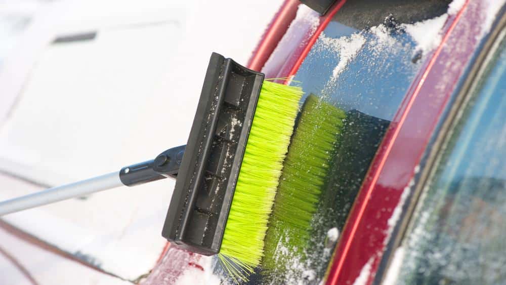 A man is cleaning snow with a brush from his car window.